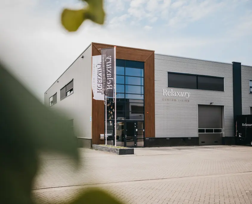 Modern industrieel gebouw met grote glazen ingang, houten en metalen accenten aan de buitenkant en Relaxury-borden aan de voorkant. Buiten hangen twee verticale vlaggen; op de voorgrond is gedeeltelijk een bladplant te zien.