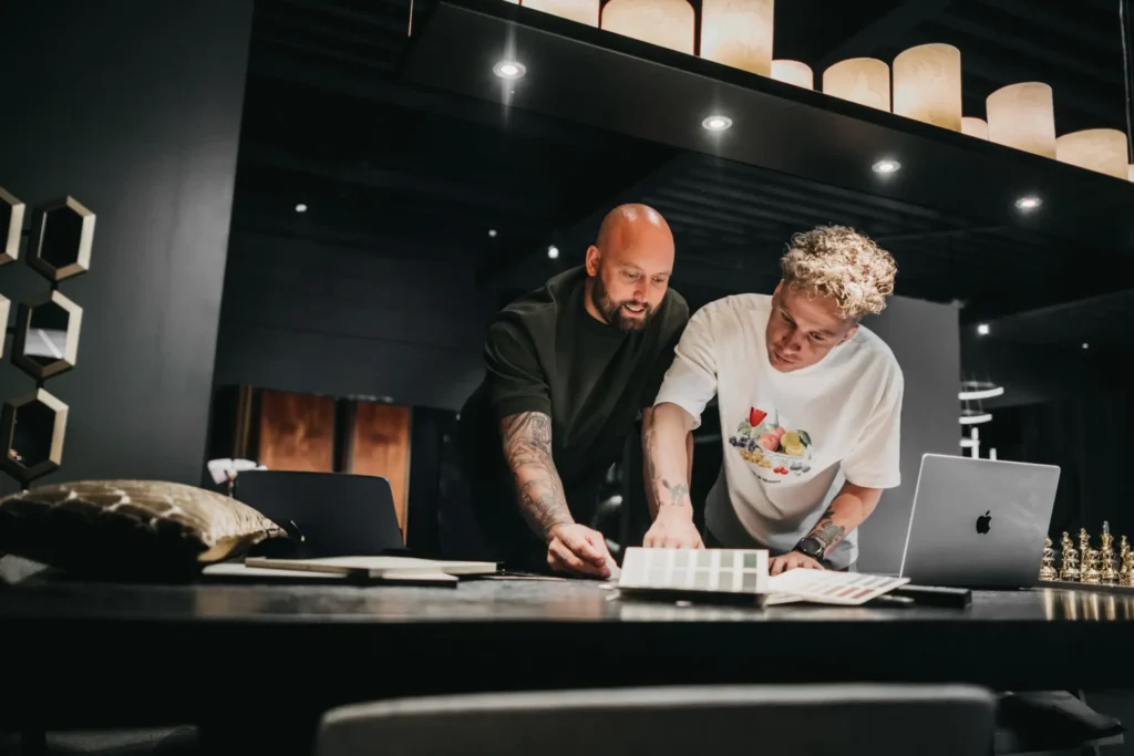 Twee mensen staan aan een bureau en bekijken samen ontwerpvoorbeelden en documenten. Ze lijken samen te werken in een moderne, schemerig verlichte werkruimte met een laptop en een schaakspel op tafel.