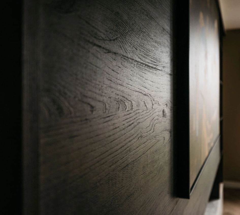 Close-up van een donker houten oppervlak met zichtbare nerfpatronen en textuur, met een deel van een ingelijst kunstwerk dat rechts aan de muur hangt. De verlichting creëert een subtiele glans op het hout.