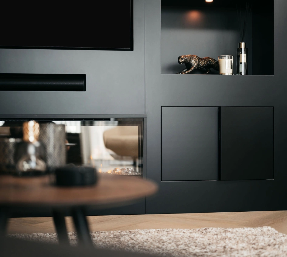 Een moderne woonkamer is voorzien van donkere ingebouwde rekken met decoratieve voorwerpen, waaronder een kaars en een klein panterbeeldje. Een houten salontafel en een zacht beige vloerkleed zijn zichtbaar op de voorgrond.