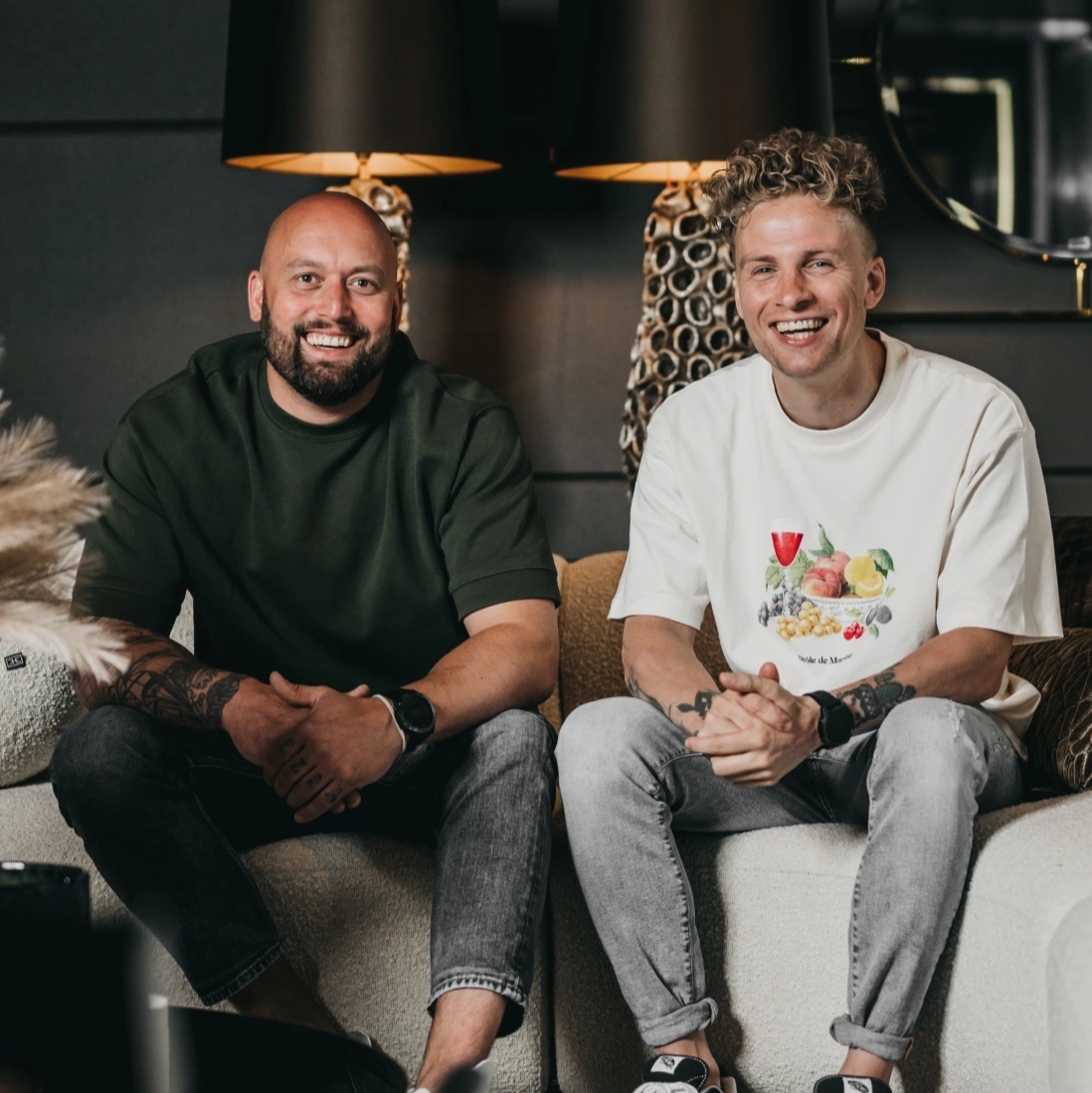 Twee mannen zitten op een bank en lachen naar de camera. De ene heeft een geschoren hoofd en baard en draagt een donkergroen shirt; de andere heeft krullend blond haar en draagt een wit grafisch t-shirt. Achter hen staan lampen en decorstukken.
