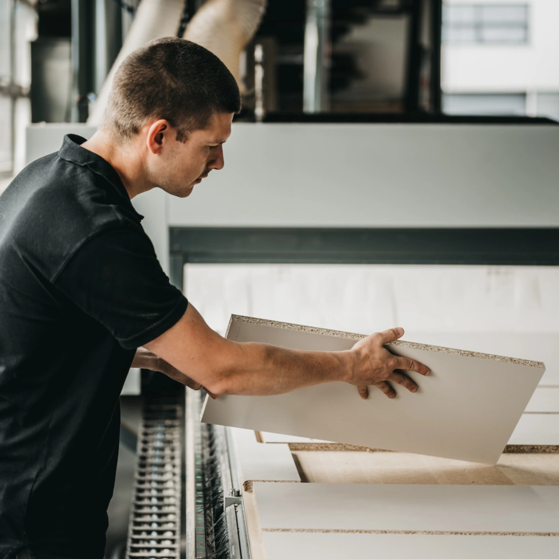 Een man in een zwart shirt plaatst een rechthoekig stuk spaanplaat op een transportband in een fabriek of werkplaats.