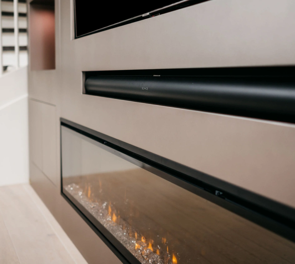 Een close-up van een moderne elektrische wandhaard met een glazen voorkant onder een strakke zwarte soundbar, geplaatst in een minimalistische beige muur. De haard heeft gloeiende sintels en een zichtbaar vlameffect.