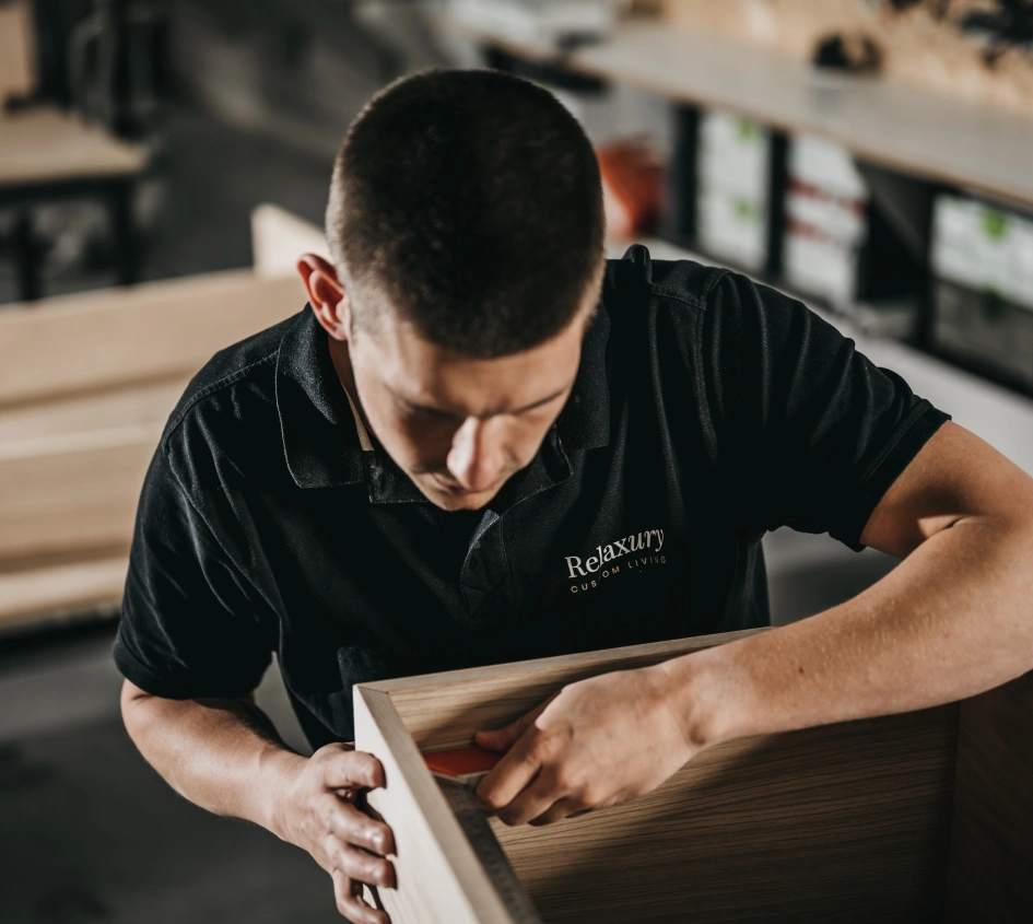 Een persoon in een Relaxury Custom Living shirt zet een houten meubel in elkaar, waarbij hij zich concentreert op het uitlijnen van de hoeken in een werkplaats.
