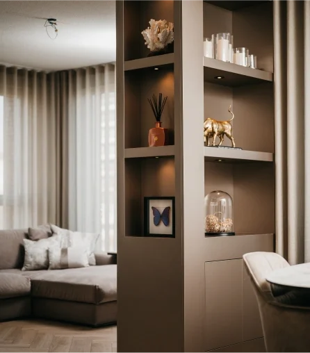 Een moderne woonkamer met een beige sofa en transparante gordijnen. Op een ingebouwde plank staan decoratieve voorwerpen zoals koraal, boeken, een gouden stier, een vlinderfoto en een glazen stolp met natuurlijke voorwerpen.