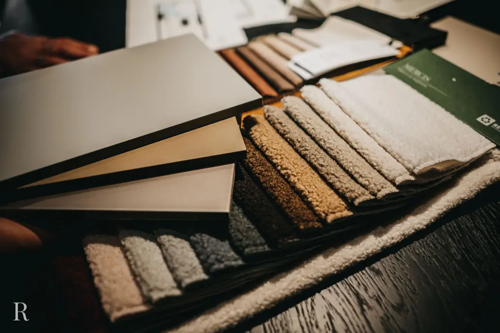 Een selectie neutraal gekleurde verf- of laminaatstalen wordt gehouden boven een display met gestructureerde stofstalen in verschillende aardetinten, gerangschikt op een tafel voor interieurontwerp.