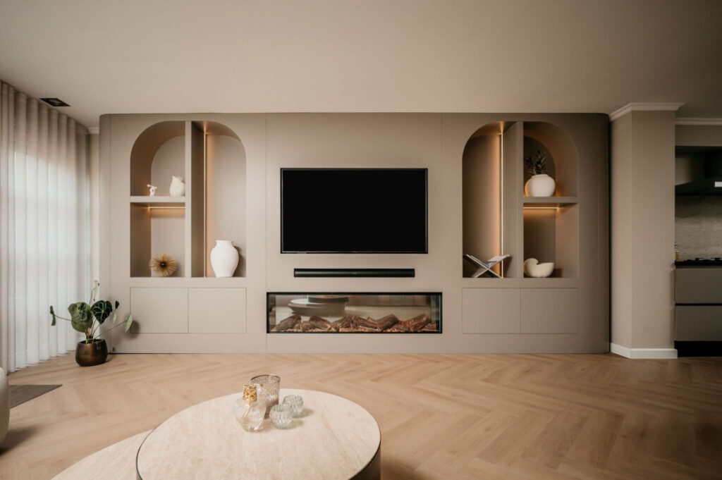 Moderne woonkamer met lichthouten visgraatvloer, ingebouwde rekken met vazen en decor, een tv aan de muur, elektrische open haard en een ronde beige salontafel met glazen voorwerpen op de voorgrond.