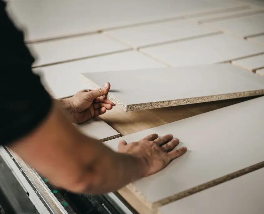 De handen van een persoon die een rechthoekig stuk spaanplaat met een wit oppervlak op een werkbank schikt of inspecteert, omringd door gelijkaardige platen.