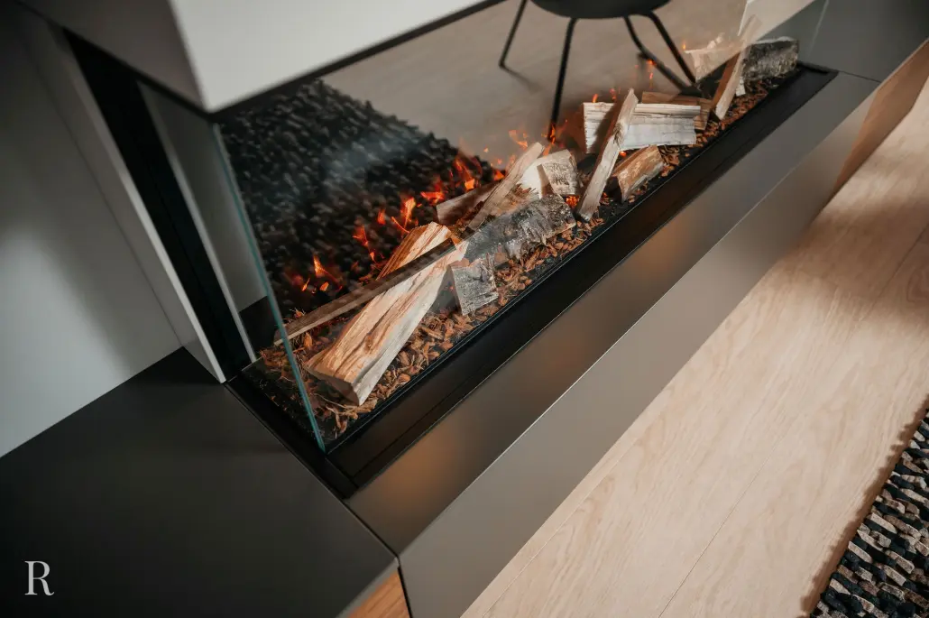 Close-up van een moderne open haard in glas met brandende houtblokken op een lichte houten vloer naast een getextureerd tapijt. De warme vlammen en strakke lijnen creëren een gezellige, eigentijdse sfeer.