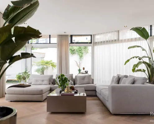 Moderne woonkamer met grote ramen, transparante gordijnen, lichtgrijze sofa's, een vierkante salontafel, een houten vloer in visgraatmotief en verschillende groene potplanten die een lichte, luchtige sfeer creëren.