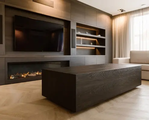 Een moderne woonkamer met een grote salontafel van donker hout, een ingebouwde open haard onder een tv aan de muur, open planken en gordijnen van vloer tot plafond naast een lichtgekleurde bank.