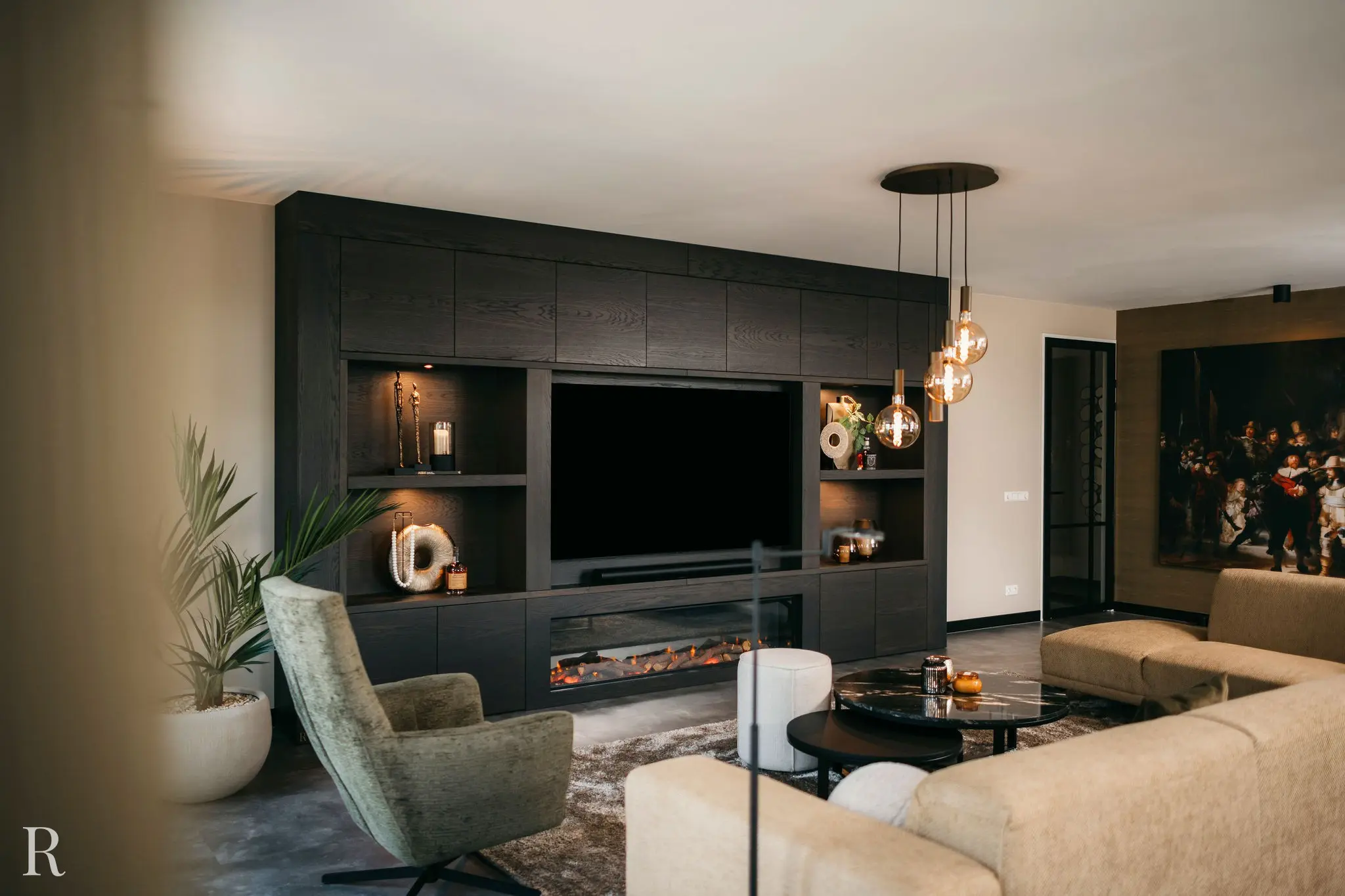 Moderne woonkamer met een donkerhouten inbouwkast met planken, decoratiespullen en een elektrische open haard. Neutrale banken, een groene fauteuil, een ronde salontafel en hangende hanglampen maken de gezellige ruimte compleet.