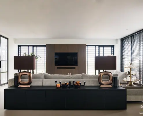 Moderne woonkamer met een neutrale bank, zwart dressoir, twee koperen lampen, decoratieve items, grote ramen met jaloezieën en een ingebouwd tv-meubel tegen een houten accentmuur. De ruimte is licht en minimalistisch.