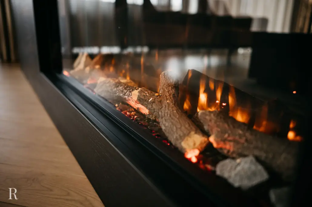 Een close-up van een moderne elektrische open haard met realistische kunststammen en gloeiende sintels, geplaatst in een strak zwart frame op een houten vloer in een gezellige omgeving binnenshuis.