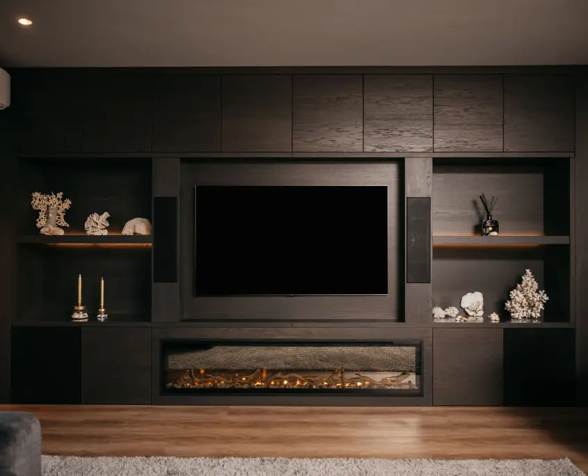 Een moderne woonkamer met een donkerhouten entertainmentmeubel met een grote tv, ingebouwde planken met decoratieve voorwerpen, kaarsen en wit koraal, en een elektrische open haard eronder. De vloer is van hout en de verlichting is warm.