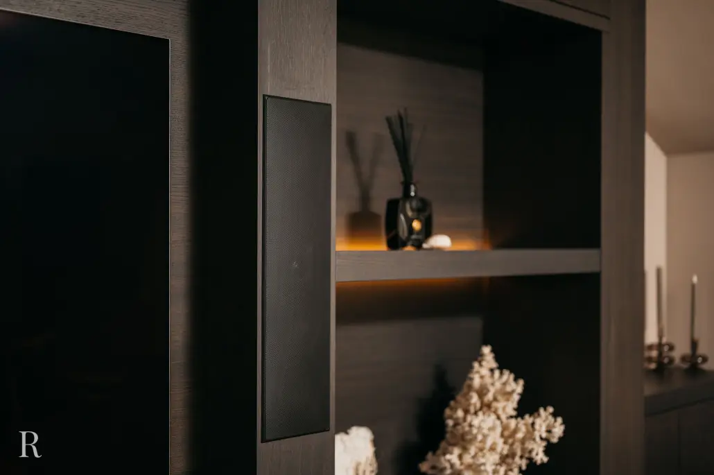 Een modern donkerhouten rek met decoratieve voorwerpen, waaronder een koraalsculptuur, kaarsen en een rietverdamper, zacht verlicht door ingebouwde verlichting. Een tv-scherm en luidspreker zijn gedeeltelijk zichtbaar aan de linkerkant.