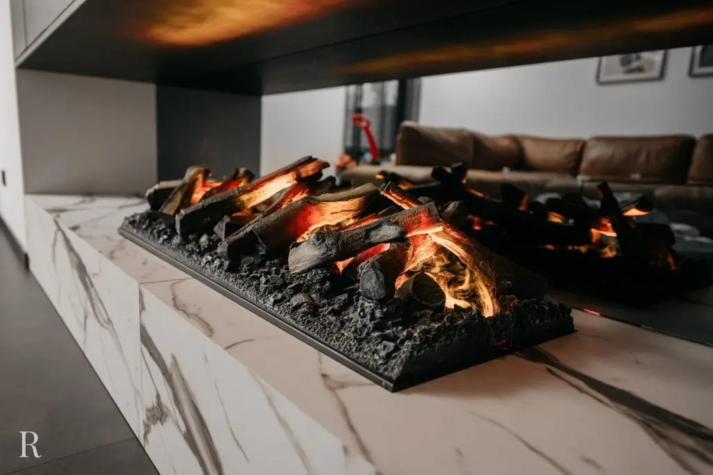 Moderne elektrische haard met realistische kunststammen en gloeiende sintels op een wit marmeren schouw in een stijlvolle woonkamer met een bruine bank op de achtergrond.