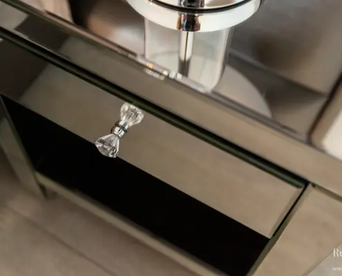 Een close-up van een spiegelend badmeubel met een ladeknop van helder glas en een chromen kraan erboven, die delen van de kamer weerspiegelt.