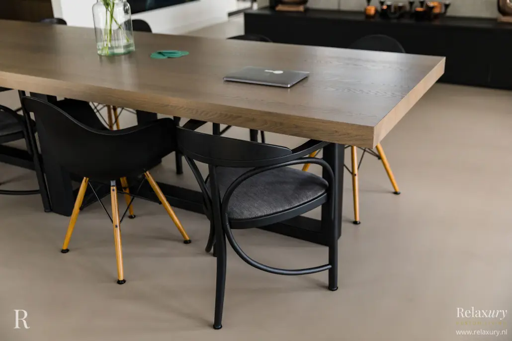 Een moderne eettafel met een houten blad, omringd door zwarte stoelen met gouden poten, staat op een beige vloer. Een gesloten laptop en een bril liggen op tafel. Het Relaxury-logo verschijnt in de rechterbenedenhoek.