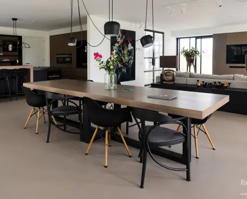 Moderne eethoek met een lange houten tafel, zes zwarte stoelen, hanglampen boven en een vaas met bloemen. De open ruimte omvat een keuken en een leefruimte met een minimalistische inrichting en neutrale tinten.