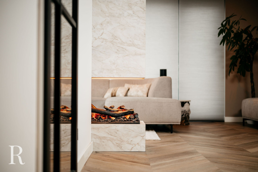 Elegante woonkamer met een marmeren open haard met brandende houtblokken, visgraat houten vloeren, een neutrale bank met kussens en een kamerplant op de achtergrond.