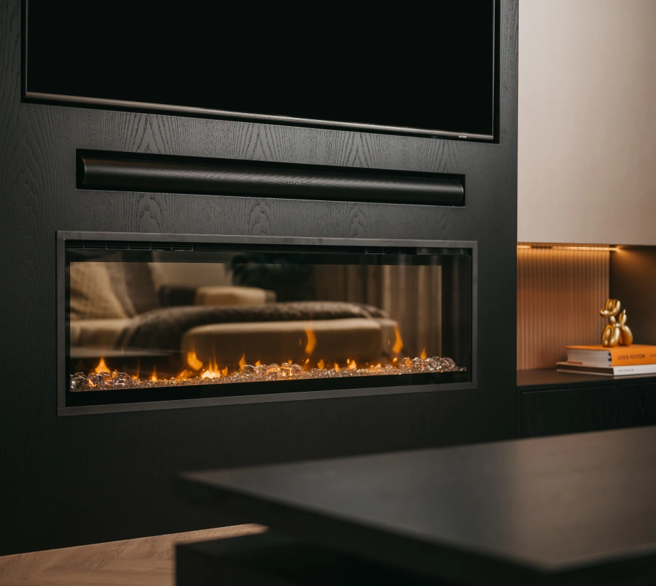 Moderne woonkamer met een lange rechthoekige elektrische open haard ingebouwd in een strakke zwarte muur, vlammen gloeien achter glas; een bank en gezellige inrichting worden weerspiegeld in het oppervlak van de open haard.