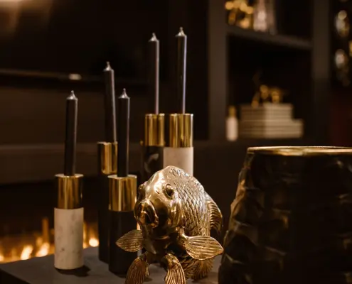 Een decoratieve display met gouden en witte kandelaars met zwarte kaarsen, een glanzend gouden visbeeldje en een donkere vaas met textuur, tegen een zacht verlichte achtergrond met een warme gloed.