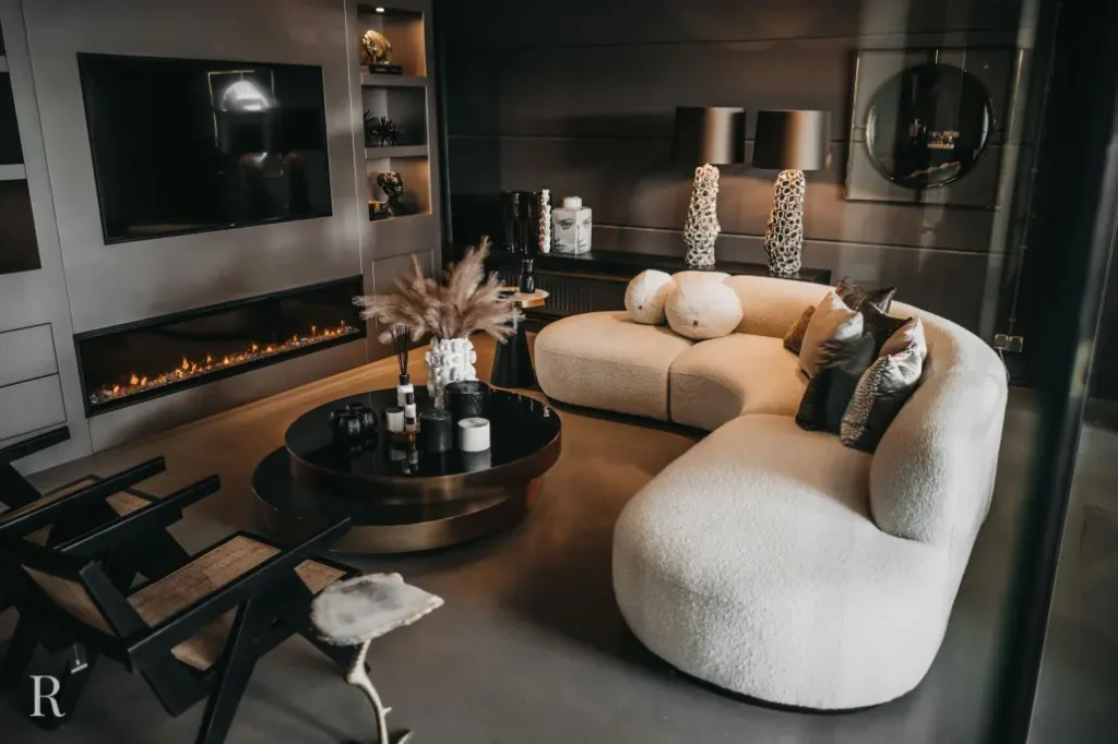 Een moderne woonkamer met een gebogen beige bank, een ronde zwarte salontafel versierd met vazen en pampagras, zwarte accentstoelen, een tv aan de muur, een lineaire open haard en twee lampen met patronen op een donkere console.