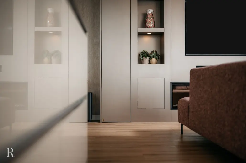 Moderne woonkamer met lichthouten vloeren, ingebouwde planken met een vaas en potplanten, een bruine bank en een flatscreentelevisie die gedeeltelijk zichtbaar is aan de muur. De scène wordt weerspiegeld in een glanzend oppervlak.