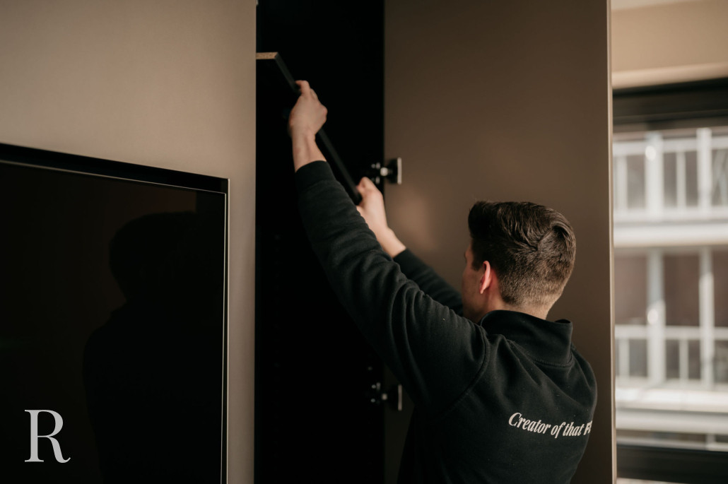 Een man in een zwarte trui zet een zwart rek in elkaar in een moderne kamer, bij een groot raam en een flatscreen-tv.