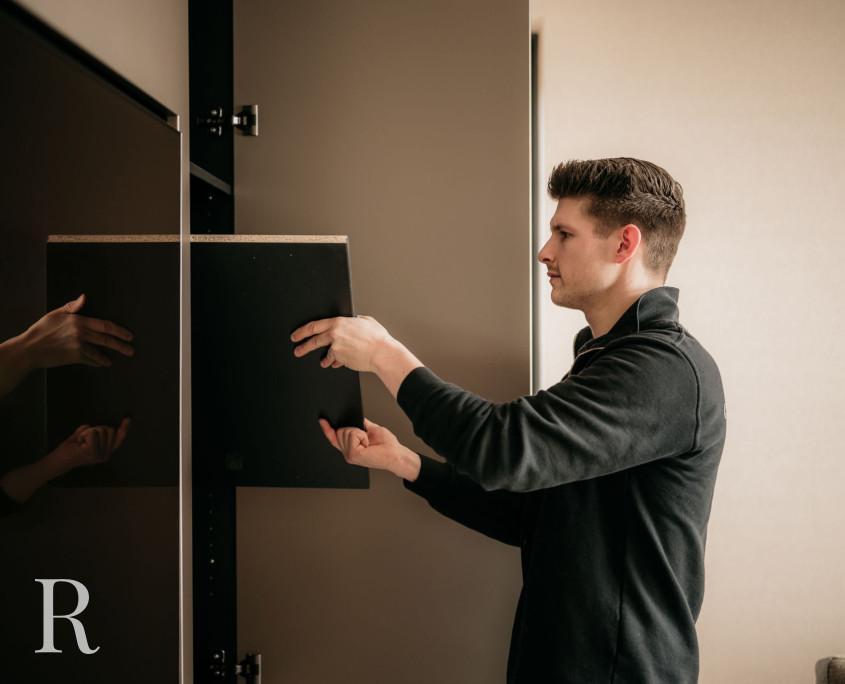 Een man in een zwarte jas installeert of past een zwarte plank aan in een kast. Hij kijkt naar de kast en gebruikt beide handen om de plank op zijn plaats te krijgen. Linksonder staat een wit R-logo.