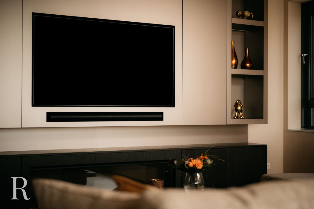 Een moderne woonkamer met een grote flatscreentelevisie aan een beige muur, planken met decoratieve gouden vazen en een beeldje, een donkere mediaconsole en een glazen vaas met oranje bloemen op een tafel.