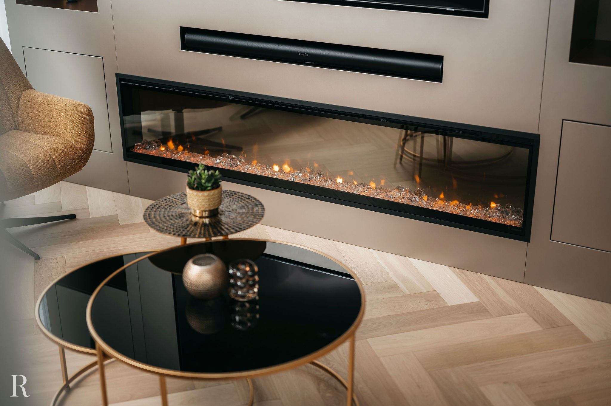 Een moderne woonkamer met een lange, ingebouwde elektrische open haard onder een soundbar, een ronde beige stoel en twee zwartglazen salontafels met gouden frames, op een ervan staan een vetplant in pot en decoratieve voorwerpen.