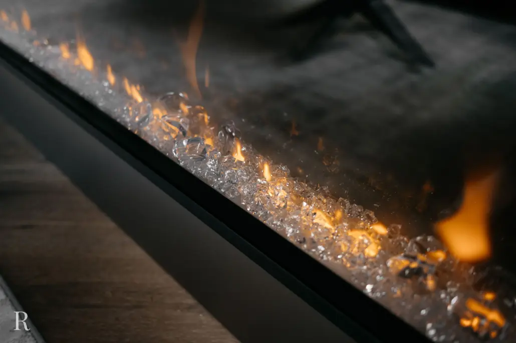 Close-up van een moderne elektrische open haard met oranje vlammen en kristallen van helder glas, in een strak zwart frame op een houten vloer.
