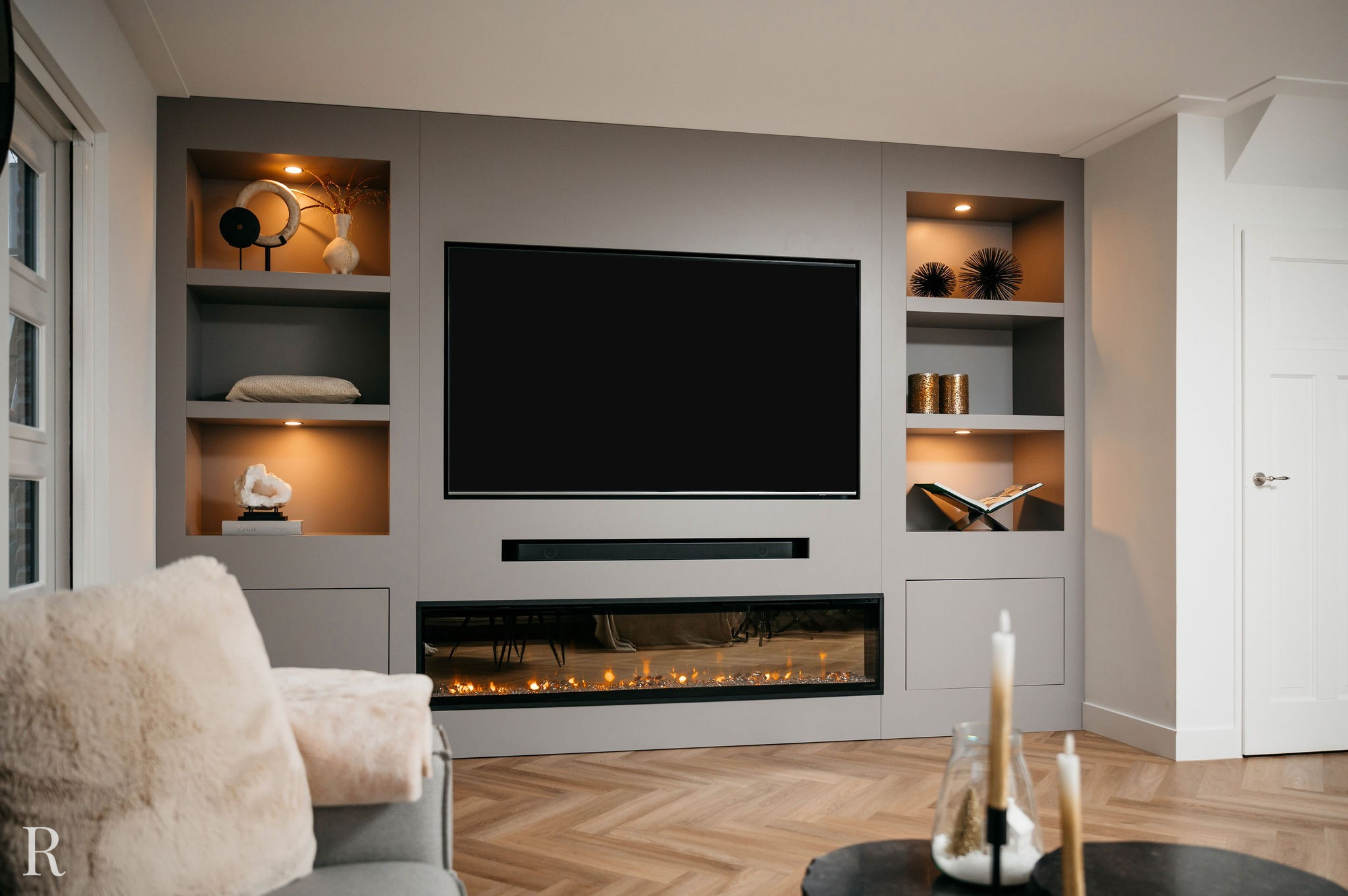 Een moderne woonkamer met een ingebouwd entertainmentcentrum met een grote tv, planken met decoratieve items, een lange elektrische open haard eronder en gezellige meubels op de voorgrond. Warme verlichting creëert een comfortabele sfeer.