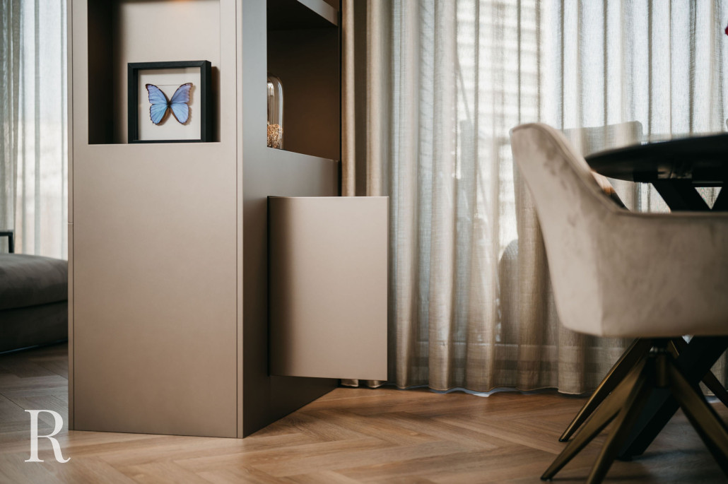 Een moderne leefruimte met een gedeeltelijk uitzicht op een beige kast met een open kastdeur, een ingelijste blauwe vlinder, een glazen koepel, een lichte fauteuil en gordijnen van vloer tot plafond die natuurlijk licht binnenlaten.