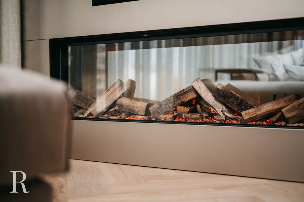Een moderne, glazen open haard met gestapelde houten houtblokken en gloeiende sintels staat in een stijlvolle kamer met lichte houten vloeren en zacht, neutraal gekleurd meubilair op de achtergrond.