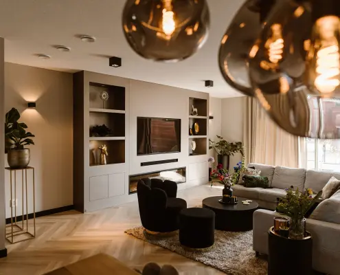 Moderne woonkamer met neutrale tinten, een grote grijze sofa, ronde zwarte salontafel, ingebouwde planken, flatscreentelevisie, planten, zachte verlichting en decoratieve hanglampen op de voorgrond.