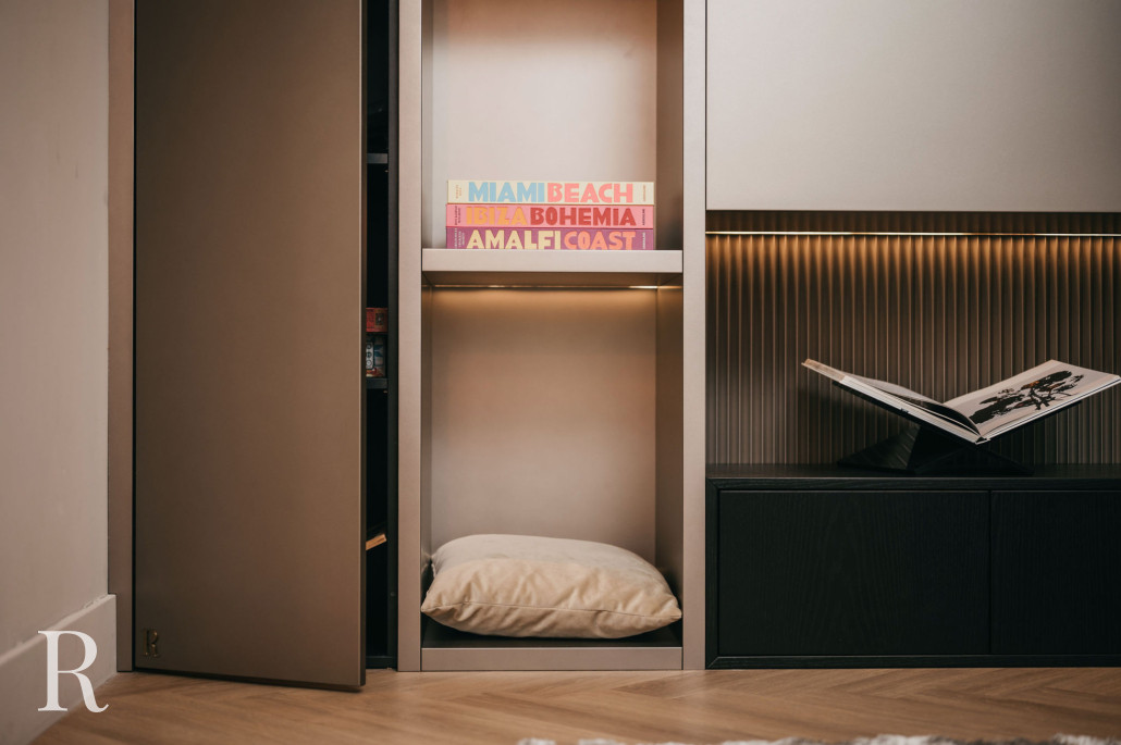 Op een ingebouwde plank staan kleurrijke reisboeken, een lichtgekleurd kussen op een lagere plank en een open boek op een donkere kast. Zachte verlichting creëert een gezellige sfeer.