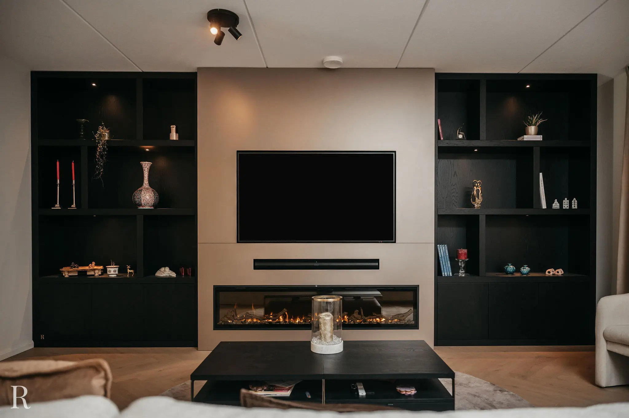Moderne woonkamer met een tv aan de muur boven een elektrische open haard, geflankeerd door zwarte rekken met kaarsen, vazen, boeken en decoratieve voorwerpen; op de voorgrond staat een salontafel.