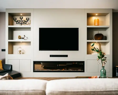 Een moderne woonkamer met een ingebouwd entertainmentcentrum met een flatscreentelevisie, decoratieve planken met kunst en vazen en een strakke elektrische open haard eronder. Een bank en een salontafel met een vaas bloemen staan op de voorgrond.