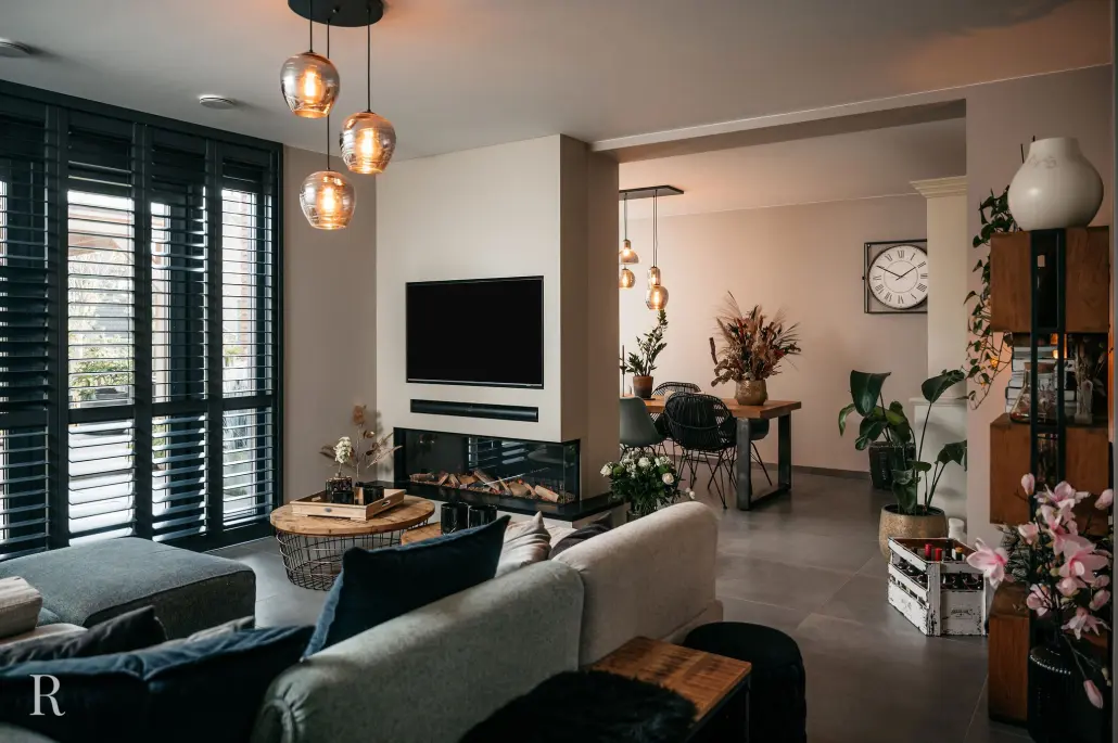 Moderne woonkamer met neutrale tinten, een tv aan de muur boven een open haard, grote ramen met donkere luiken, hanglampen, een gezellige bank en een eethoek op de achtergrond met planten en een grote klok aan de muur.