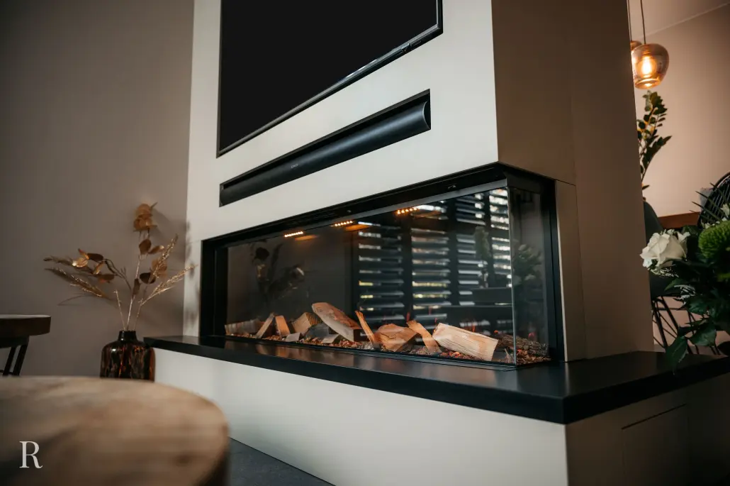 Moderne binnenhaard met glazen zijkanten, gevuld met decoratieve houtblokken. Het omringende decor bestaat uit gedroogde planten, een houten kruk en groen gebladerte in een goed verlichte, eigentijdse kamer.