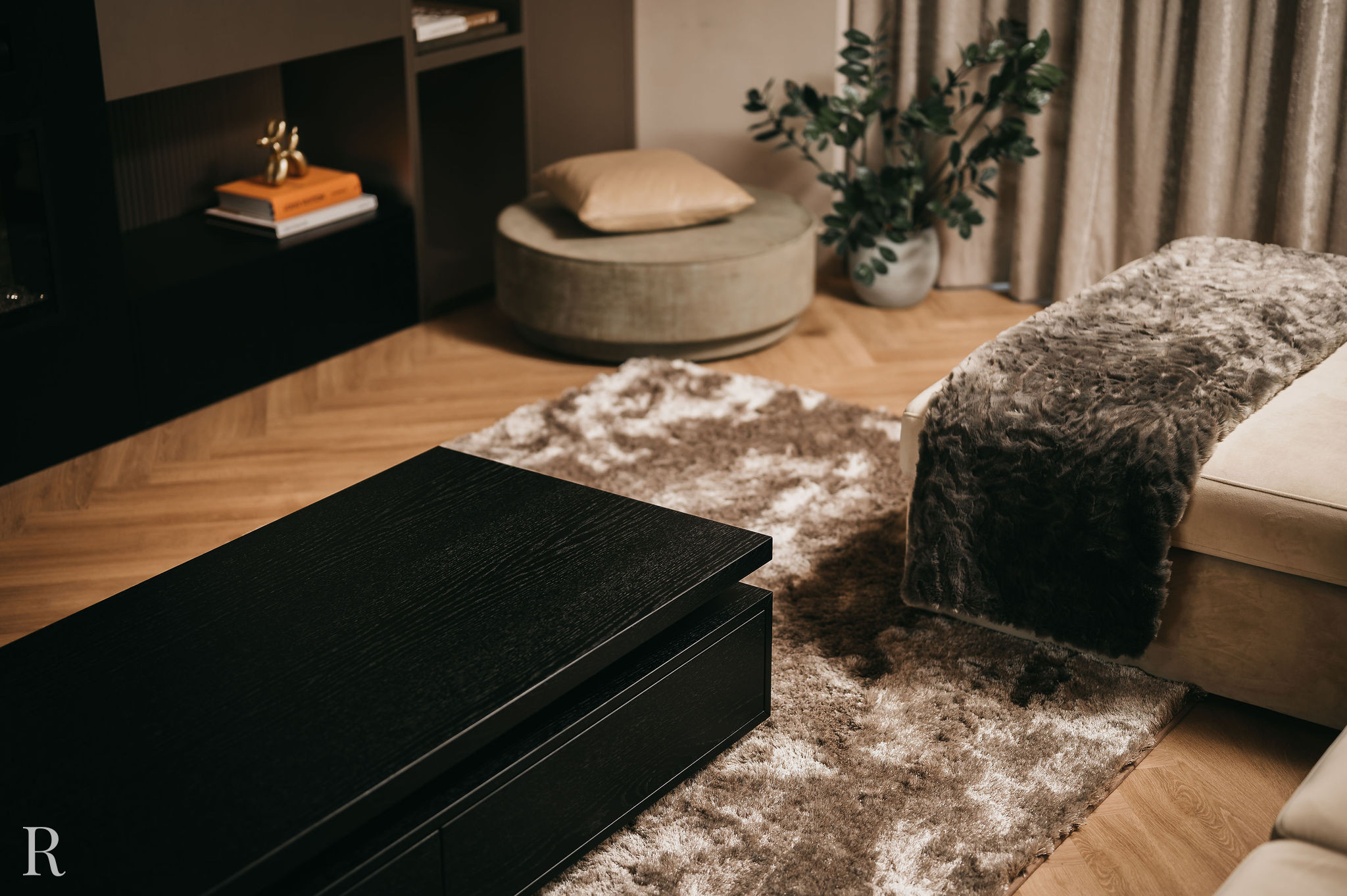 Een gezellige woonkamer met een donkerhouten salontafel, een beige bank met een kleed van grijs bont, een rond voetenbankje met kussen, een pluchen vloerkleed, een plant en planken met boeken en decoratieve voorwerpen.