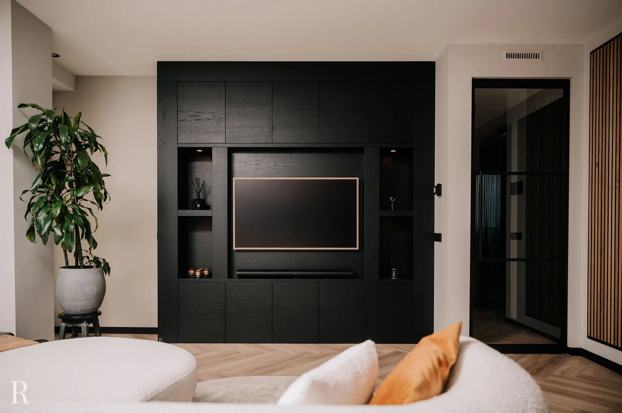 Moderne woonkamer met een zwart ingebouwd tv-meubel, flatscreen tv, soundbar, planken met decor, grote potplant aan de linkerkant, een witte bank met oranje en witte kussens en houtaccenten.