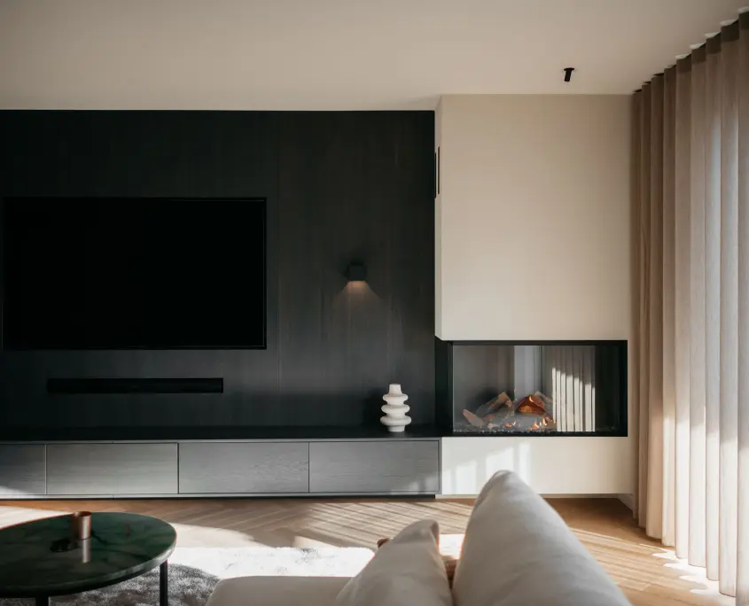 Moderne woonkamer met een grote tv aan de muur, donkerhouten lambrisering, minimalistische kasten, een ingebouwde glazen open haard met houtblokken, beige gordijnen en een deel van een witte bank op de voorgrond. Natuurlijk licht vult de ruimte.