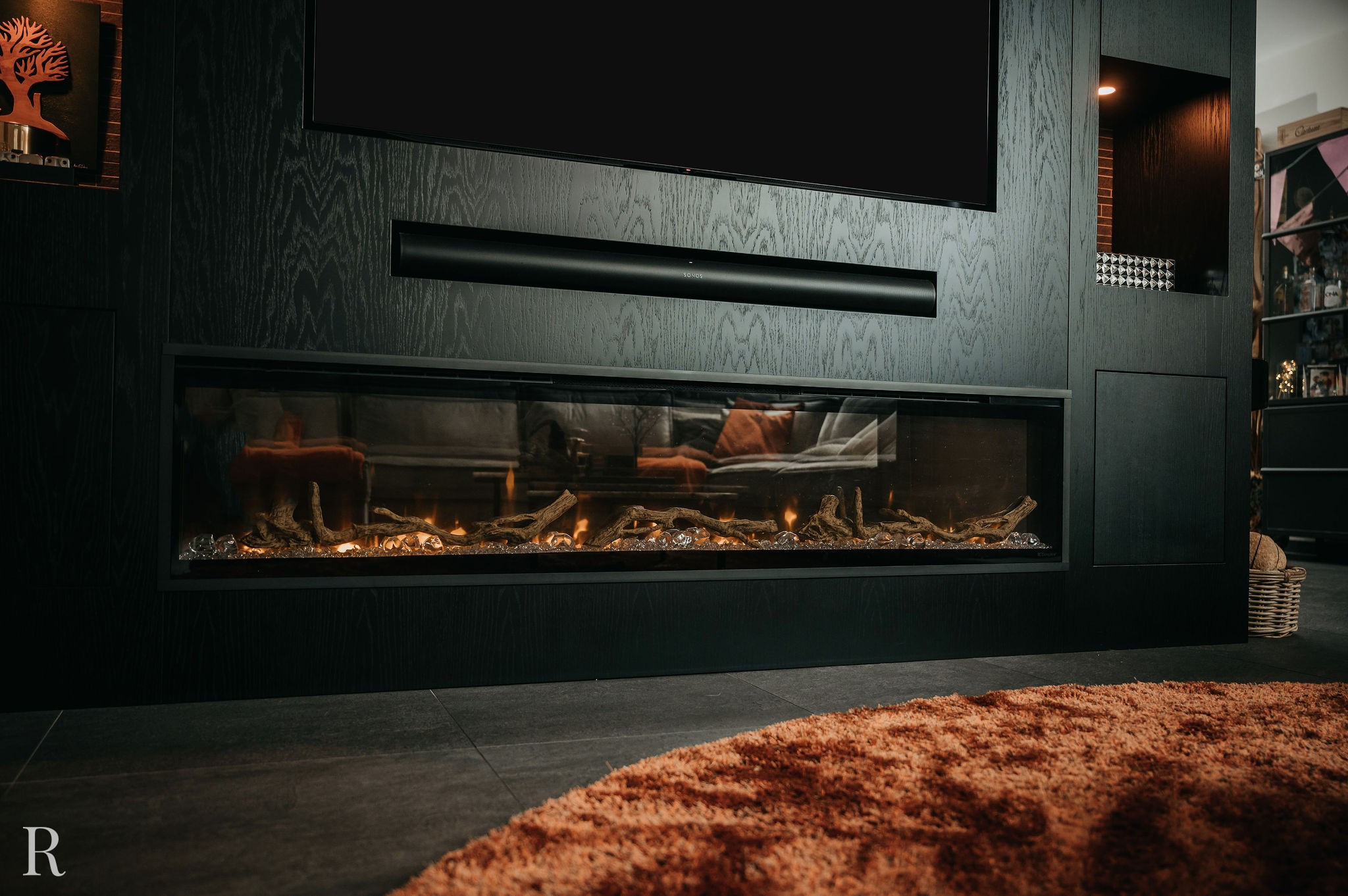 Een moderne zwarte wandhaard straalt een warme gloed uit, met houtblokken en sintels erin. Erboven staat een tv en ervoor ligt een knus oranje vloerkleed met structuur op de donkere tegelvloer.