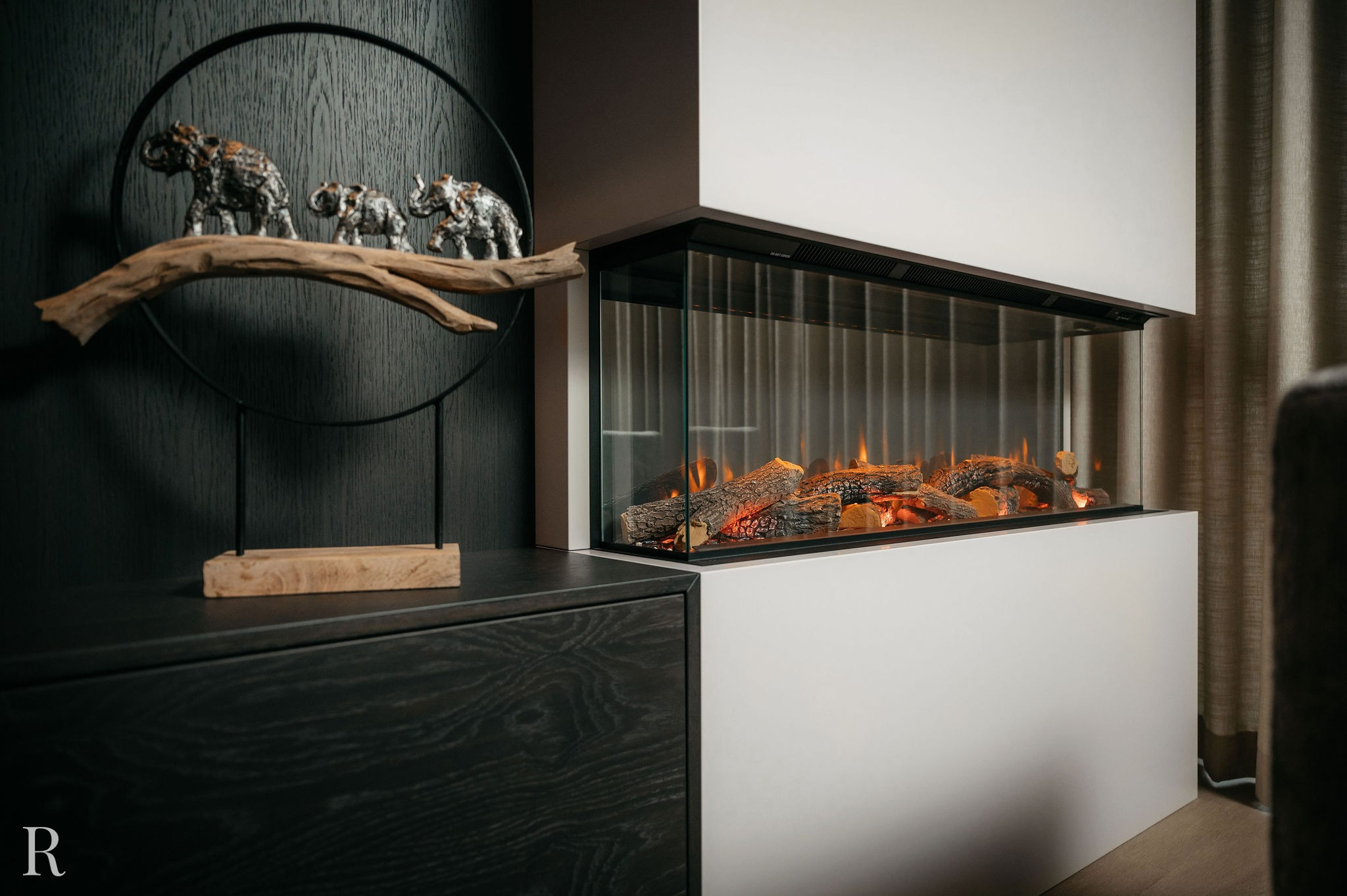 Een moderne elektrische open haard met gloeiende houtblokken is ingebouwd in een witte muur. Daarnaast staat in een donkere kast een decoratieve sculptuur van drie olifanten op een stuk drijfhout in een rond metalen frame.