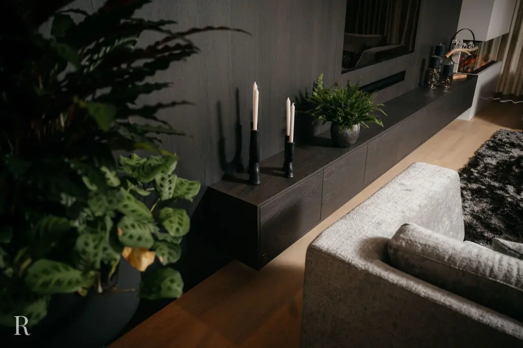 Een moderne woonkamer met een grijze bank, zwarte zwevende kast, decoratieve zwarte kandelaars met niet aangestoken kaarsen, groene potplanten en een ruig donker vloerkleed op een lichte houten vloer.