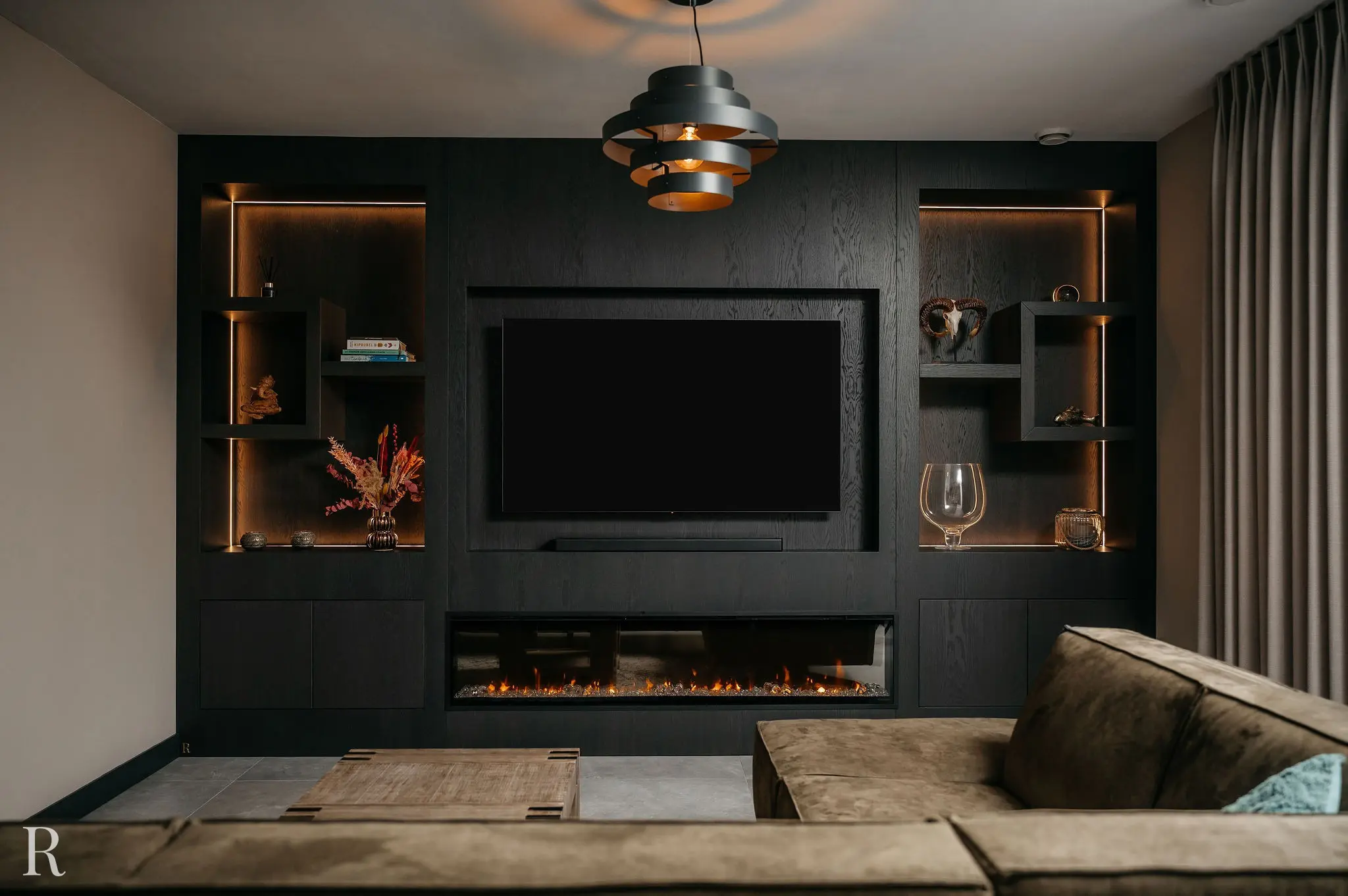 Moderne woonkamer met een zwart ingebouwd entertainmentcentrum, voorzien van een grote tv, planken met decoratieve items, een elektrische open haard en een bruine sectionele bank op de voorgrond. Warme verlichting accentueert de planken.